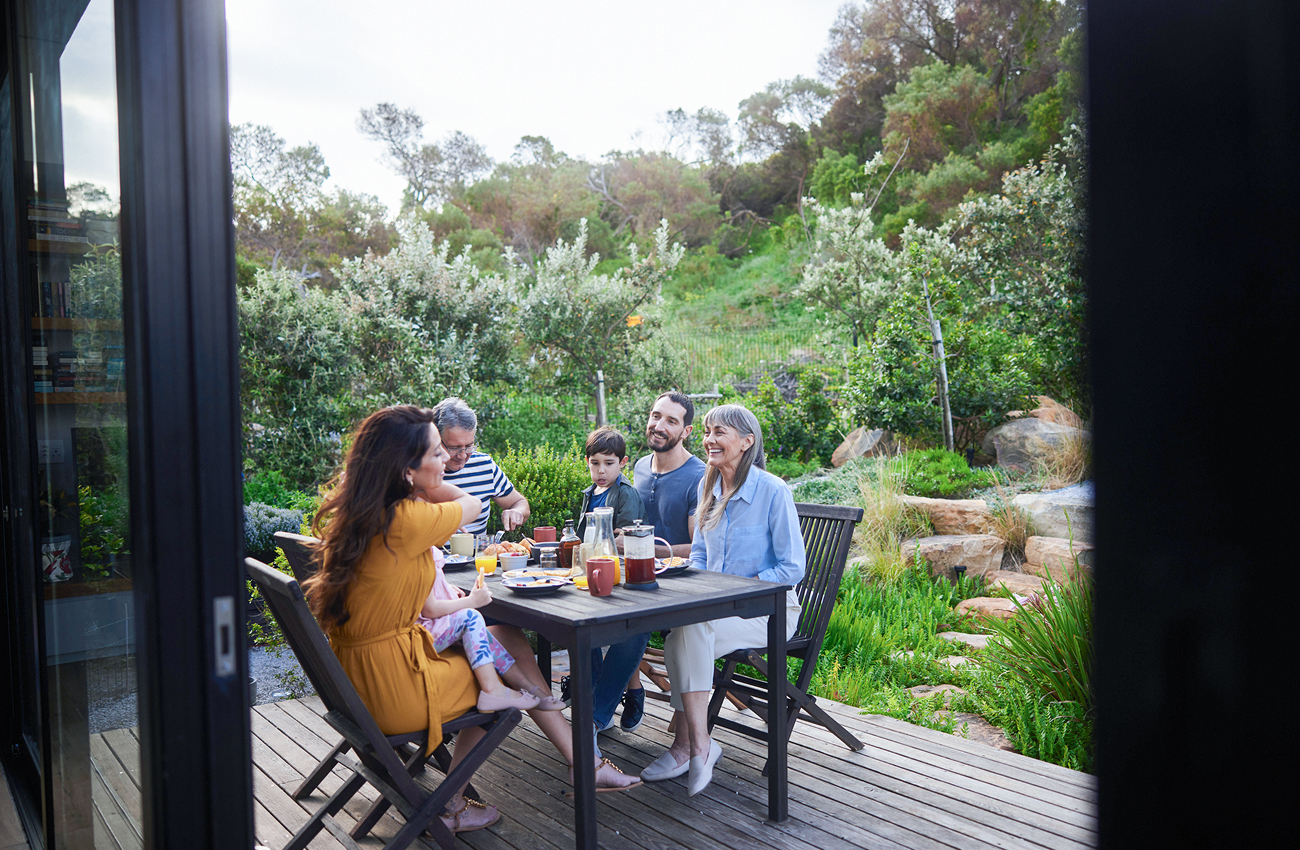 Eine fröhliche Familie versammelt sich im Freien um einen Holztisch und frühstückt gemeinsam inmitten üppigen Grüns und einer heiteren Atmosphäre.